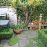 In een klein hoekje in de tuin staat een bakstenen muur bedekt met klimop, staat een ouderwetse metalen tafel met het onderstel van een naaimachine, staan stapels terracotta potten op een rand en groeien er allerlei groene planten. Dit alles wordt omgeven door een gesnoeide buxushaag.