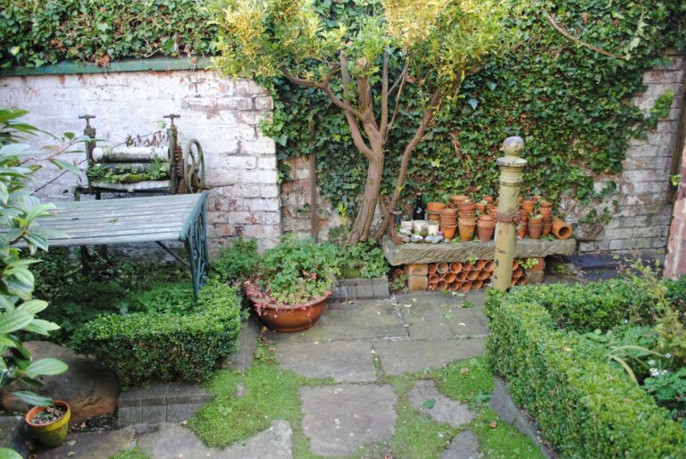 In een klein hoekje in de tuin staat een bakstenen muur bedekt met klimop, staat een ouderwetse metalen tafel met het onderstel van een naaimachine, staan stapels terracotta potten op een rand en groeien er allerlei groene planten. Dit alles wordt omgeven door een gesnoeide buxushaag.