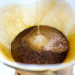 Close-up van heet water dat over gemalen koffie in een papieren filter in een pour-over koffiezetapparaat wordt gegoten. Het water creëert een bloom-effect, waardoor de koffiedik uitzet en bubbels vrijkomen terwijl het brouwproces begint.