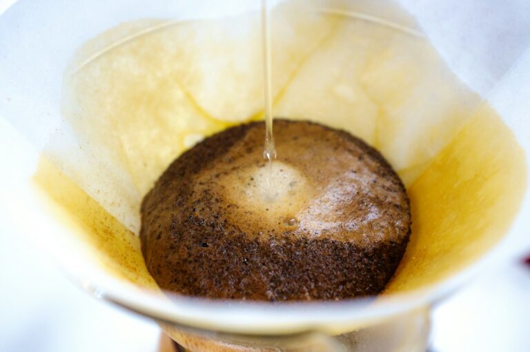 Close-up van heet water dat over gemalen koffie in een papieren filter in een pour-over koffiezetapparaat wordt gegoten. Het water creëert een bloom-effect, waardoor de koffiedik uitzet en bubbels vrijkomen terwijl het brouwproces begint.