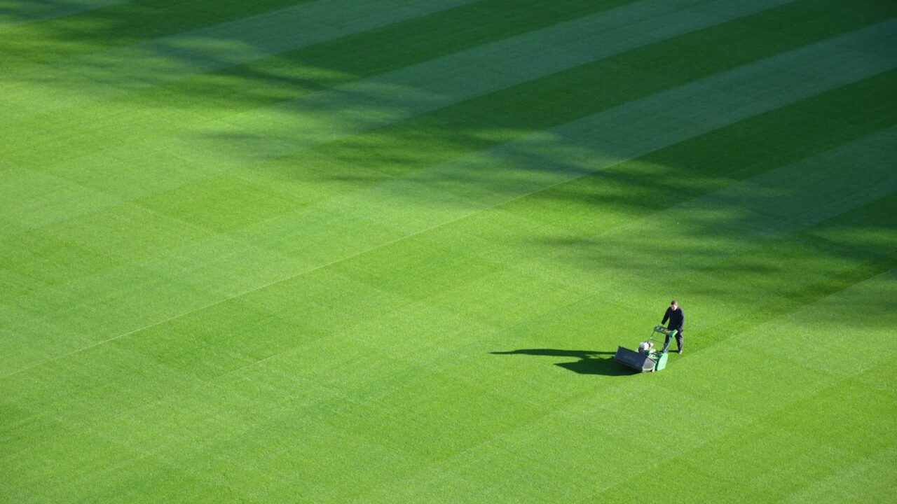 Iemand duwt een grasmaaier over een groot, netjes gestreept groen veld, waarbij schaduwen diagonale patronen op het gras creëren.