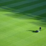 Iemand duwt een grasmaaier over een groot, netjes gestreept groen veld, waarbij schaduwen diagonale patronen op het gras creëren.