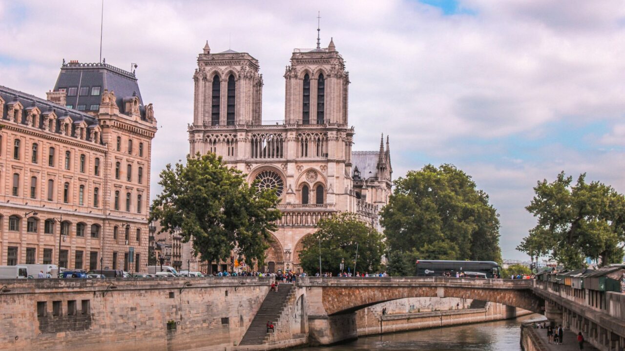 Notre Dame Parijs Een grote stenen kathedraal met twee torens staat aan een rivier, omgeven door bomen en historische gebouwen. Mensen lopen langs de rivieroever en een brug overspant het water voor de kathedraal. De lucht is gedeeltelijk bewolkt.