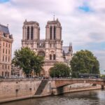 Notre Dame Parijs Een grote stenen kathedraal met twee torens staat aan een rivier, omgeven door bomen en historische gebouwen. Mensen lopen langs de rivieroever en een brug overspant het water voor de kathedraal. De lucht is gedeeltelijk bewolkt.