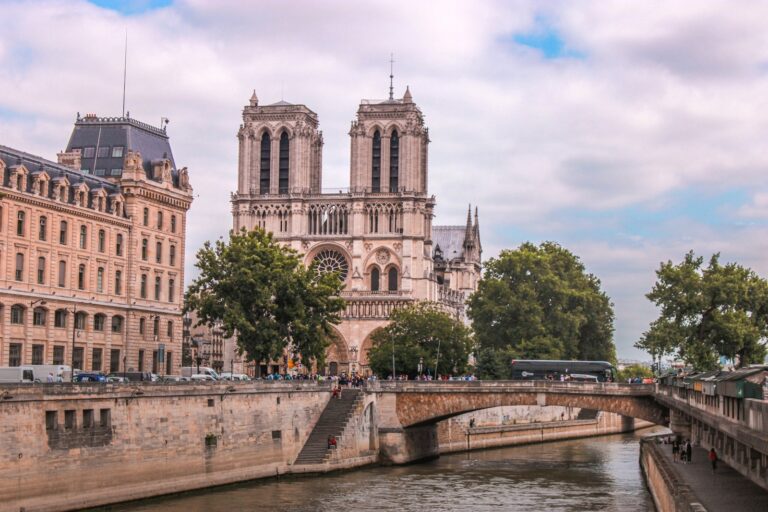 Notre Dame Parijs Een grote stenen kathedraal met twee torens staat aan een rivier, omgeven door bomen en historische gebouwen. Mensen lopen langs de rivieroever en een brug overspant het water voor de kathedraal. De lucht is gedeeltelijk bewolkt.